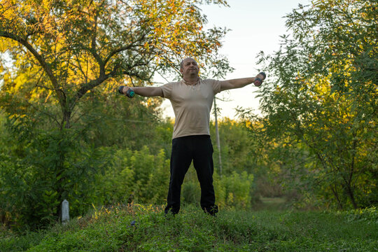 Middle aged man exercises with dumbbells held wide at shoulder height in wooded clearing. Outdoor workout shows fitness enthusiast standing tall, focused among lush greenery and autumn leaves.