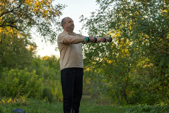 Middle aged man holds dumbbells straight ahead during outdoor workout. Exerciser stands on leafy woodland path, looking focused as he works his shoulders and arms among autumn trees.