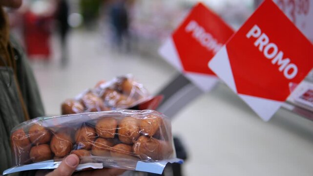 Hand holds packaged brown rolls beneath red promo signs in grocery store. Shopper compares bakery goods with marked offers, considering discounted item during routine food shopping.