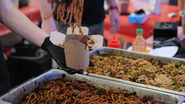 Market vendor fills open paper carton with fried insect larvae using serving tongs. Food worker portions crunchy snack beside noodle trays, presenting unusual street food to customers.