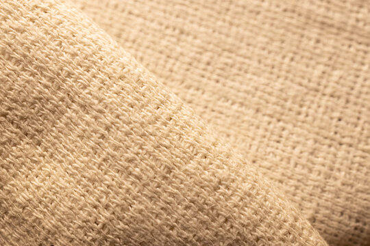 Close-up of natural beige woven fabric with a soft curved fold, visible fibers and coarse textile texture. Neutral background for design and craft.