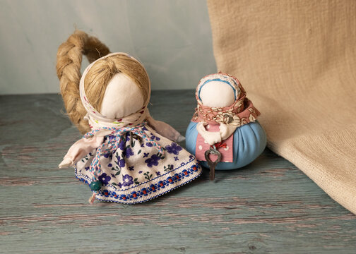 Two handmade cloth dolls in colorful patterned clothing displayed on a rustic wooden surface beside natural beige fabric. Cozy decorative still life.