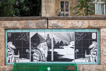 Jerusalem, Israel,  Black and white mosaic tile mural by Ephraim Moses Lilien near historic Bezalel Academy depicts Jewish men, pyramids and biblical imagery on  stone wall beside a green bench. © elenarostunova