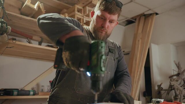 Carpenter operates green cordless drill to make precise holes into wooden blocks within workshop filled with lumber and hand tools. man wears protective gloves overalls covered in sawdust