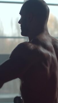 Silhouetted muscular man standing shirtless in a softly lit gym, emphasizing defined back muscles, strength, discipline, and focused athletic training