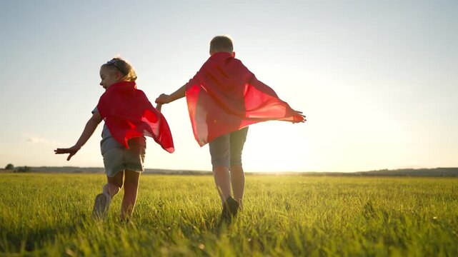 Two children wearing red superhero capes run through green field at sunset with small dog. Boy and girl holding hands while playing heroes outdoors. Kids pretending superheroes at golden hour.