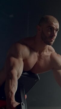 Intense shirtless bodybuilder training on exercise machine, showcasing muscular upper body strength, focus, and determination in dramatic low light gym setting