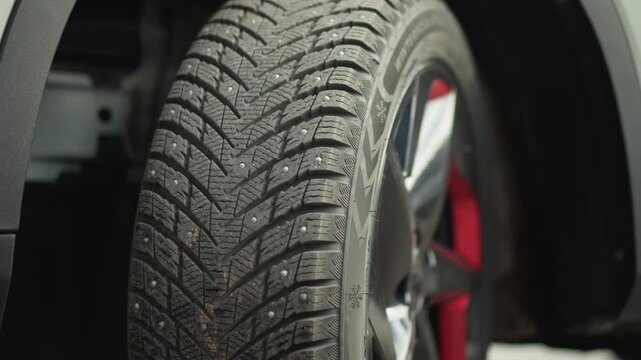 Close up on car tire showing detailed rubber treads vehicle rim and surrounding body panel. Tire features clear pattern depth and small embedded metal studs for traction. Vehicle part