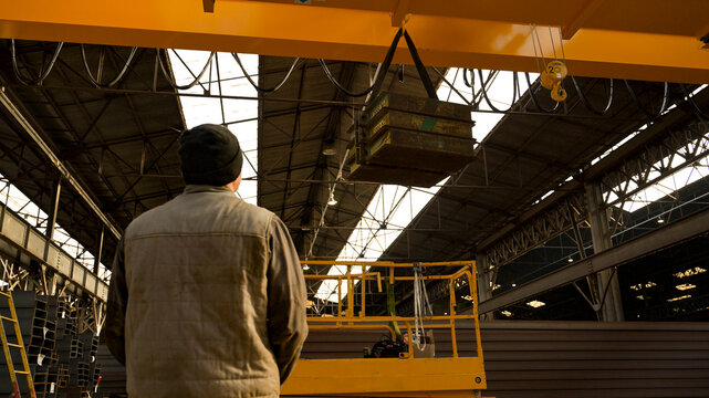 Two industrial workers handling a suspended steel load with an overhead crane in a large metal factory, showing teamwork, heavy industry and material handling.