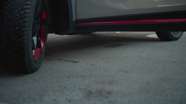 Close up view shows car wheel details on asphalt followed by black high heeled foot stepping near vehicle. Ground surface and tire treads are visible throughout