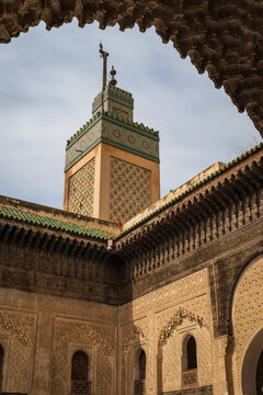 Low-angle perspective framing the ornate minaret and courtyard architecture of the historic Al-Attarine Madrasa through an intricately carved Moorish archway in Fes, Morocco