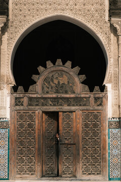 Detailed view of intricately carved wooden doors beneath a horseshoe arch and geometric stucco decorations inside the historic Al-Attarine Madrasa courtyard in Fes, Morocco