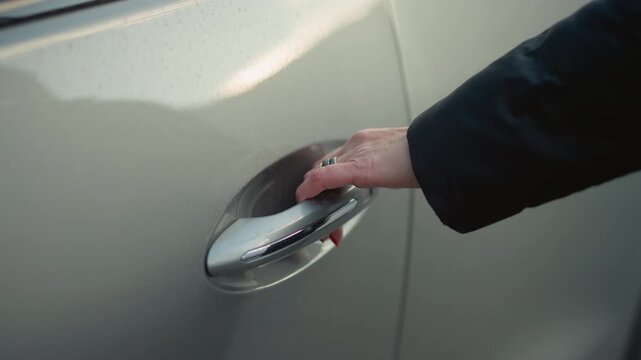 Close up view shows hand with red nail and ring reaching for silver vehicle door handle. Hand grasps pulls on lever to open car doors. Smooth metallic surface of white silver automobile body