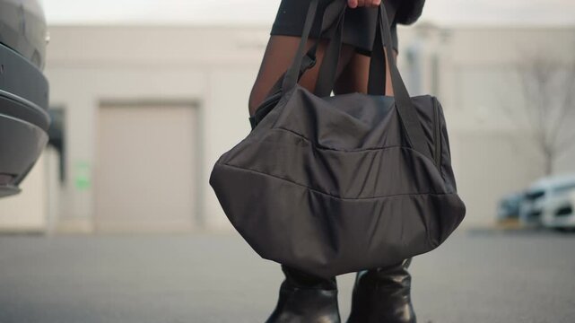 Black duffel bag held by legs in high boots with partial car and building visible. focus remains on texture detail of fabric material leather boot heel