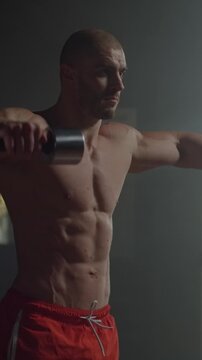 Muscular athlete performing intense dumbbell shoulder workout in dimly lit gym, showcasing defined upper body strength, focus, and disciplined fitness training
