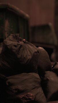 Trash bags pile up in a dimly lit room at a waste collection site. The bags are black and contain various types of waste. The smell is strong in the air.
