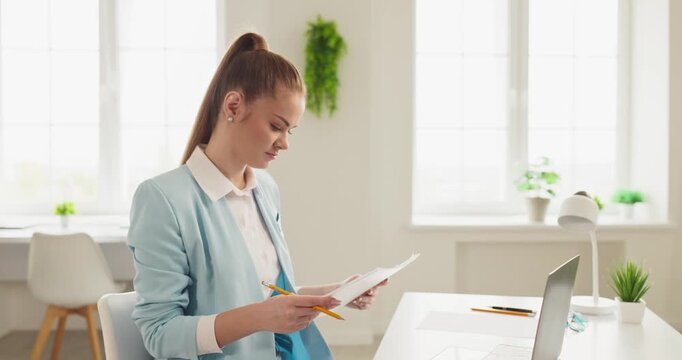 Businesswoman in office reviewing documents on laptop. Focused on analysis and planning for finance, she reviews paperwork at a tidy workplace desk. Concept of modern financial productivity