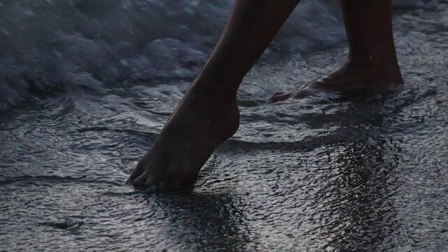 Bare feet walk through shallow tide at the shore during dusk. Extreme close-up low angle slow motion shot.