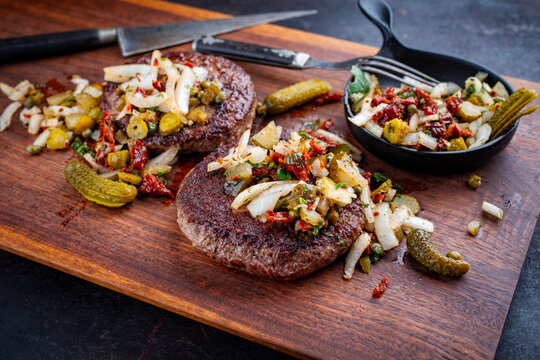 Grilled beef hamburger patties with relish made of gherkin, onions and sun dried tomatos served as close-up on a rustic wooden board