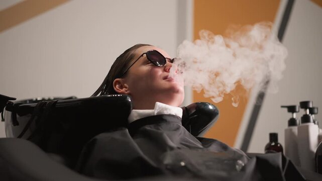 Woman reclines in salon chair with wet hair wearing sunglasses while blowing smoke. Visible elements include black cape blow dryer arm positioned near her head various bottles of products