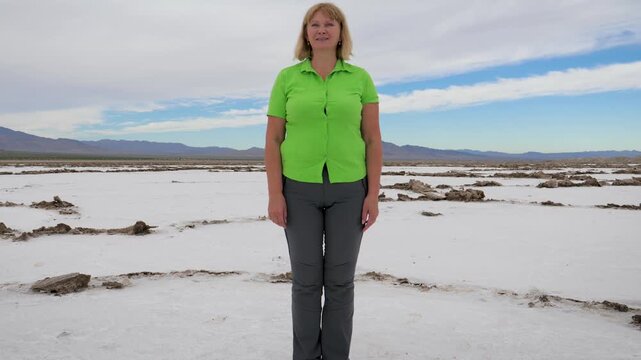 Full length portrait of female tourist standing in middle of vast, barren salt flat landscape with distant mountains under partly cloudy sky. A shot from below upward