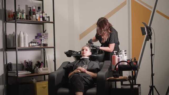Woman lies back on styling chair as another woman applies dark hair dye to her wet hair in salon. scene includes shelves with beauty products and equipment indicating ongoing hairstyling