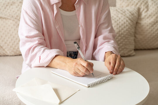 Woman in pink shirt writes with pen in spiral-bound notebook while sitting on cozy, light-colored sofa at round white coffee table. Writing personal letter in cozy, calm setting, handwritten format