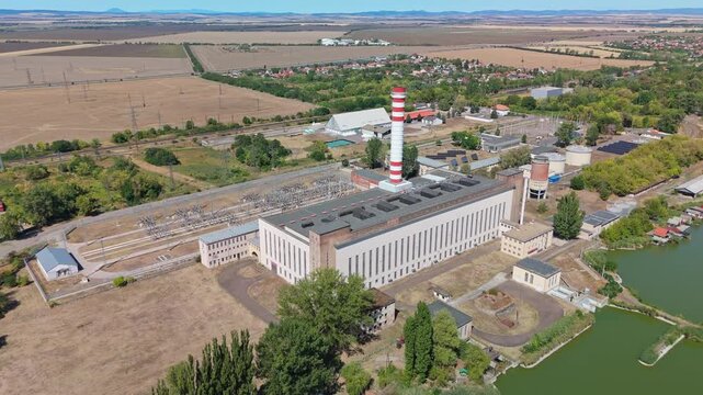 Aerial view of a thermal power plant with a striped smokestack, electrical substation, cooling lake and rail line beside farmland in Lőrinci, Hungary.