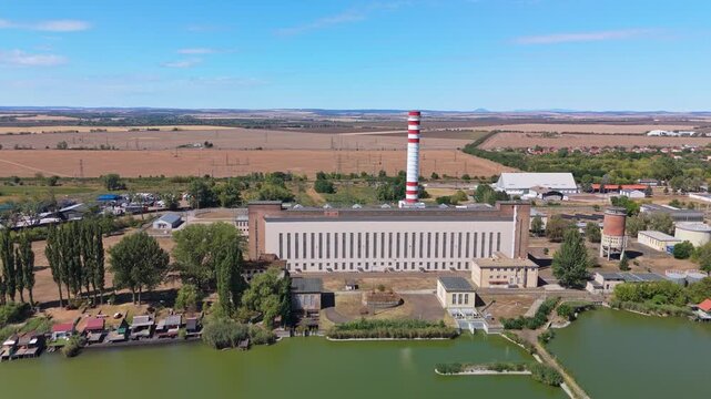 Aerial view of a thermal power plant with a striped smokestack and cooling lake, beside agricultural fields and transmission lines in Lőrinci, Hungary.