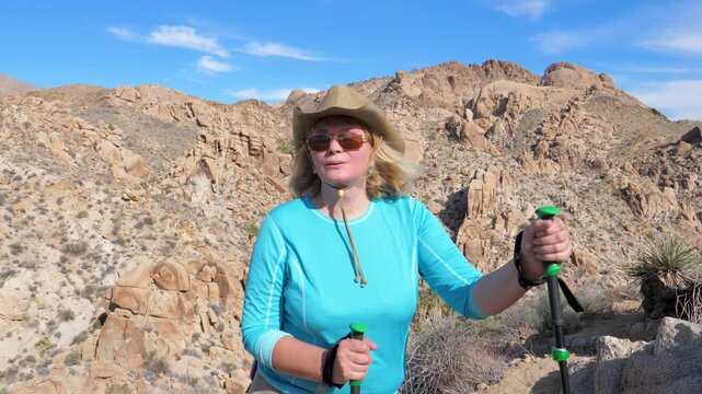 Blonde mature woman wearing hat and sunglasses is hiking with trekking poles in rocky desert canyon. Healthy outdoor lifestyle with scenic view. Female hiker is resting and taking deep breaths