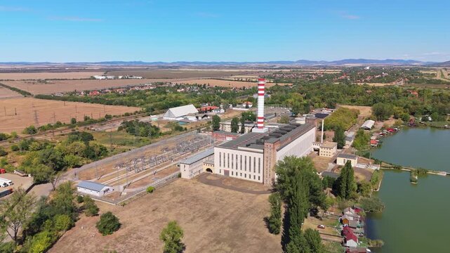 Aerial view of the M&aacute;tra Region Power Plant with a striped smokestack, electrical substation and cooling lake beside farmland in Lőrinci, Hungary.