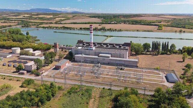 Aerial view of a thermal power plant with striped smokestack, electrical substation and cooling lake amid summer farmland and hills in Lőrinci, Hungary.