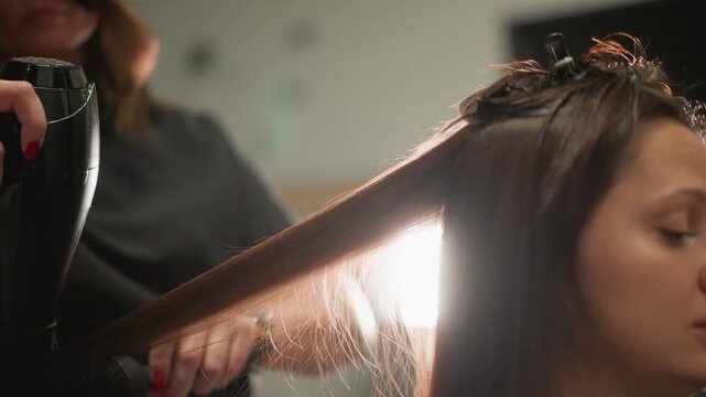 Close up shows woman receiving professional hairstyling. salon worker uses blow dryer and brush to style client wet sectioned brown hair under soft lighting