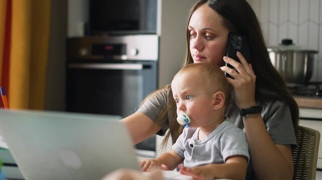 Mother working from home holds baby with pacifier on lap at desk. Busy mom types on laptop and talks on phone with infant nearby. Woman multitasks balancing remote work and childcare in kitchen.
