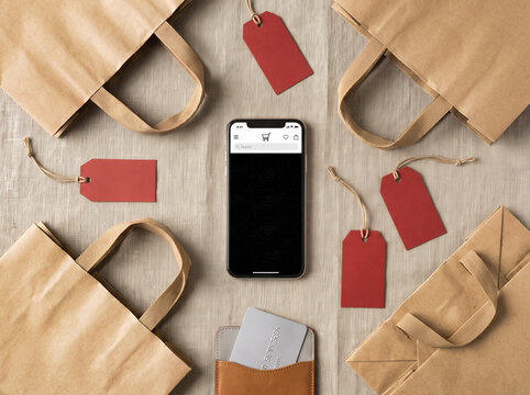 Smartphone lies on linen tablecloth alongside brown paper shopping bags, red price tags and a gray plastic credit card. Marketing promotion for Black Friday.