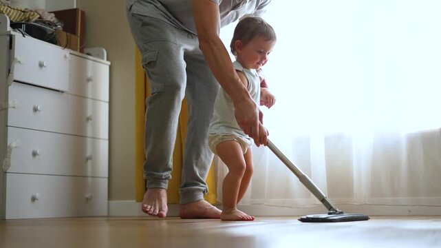 Parent helps toddler mop hardwood floor indoors. Adult guides baby hands on mop during cleaning. Toddler learns housework with parent support. Child and parent clean floor together at home.