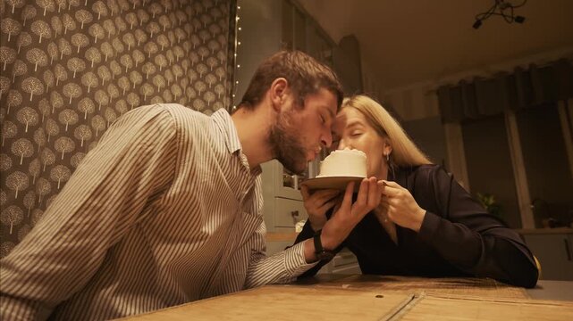 Affectionate man and woman celebrating a special occasion at home, playfully eating a small white birthday cake from the same plate and sharing a sweet kiss in a cozy kitchen at night
