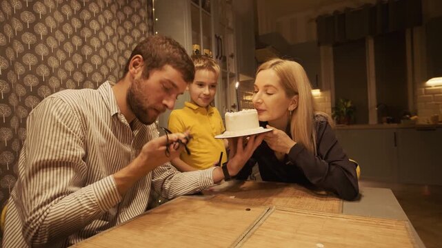 Loving family celebrating a birthday at home in the evening, with a mother holding a cake with a candle while her husband and son look on, creating a warm and happy atmosphere together