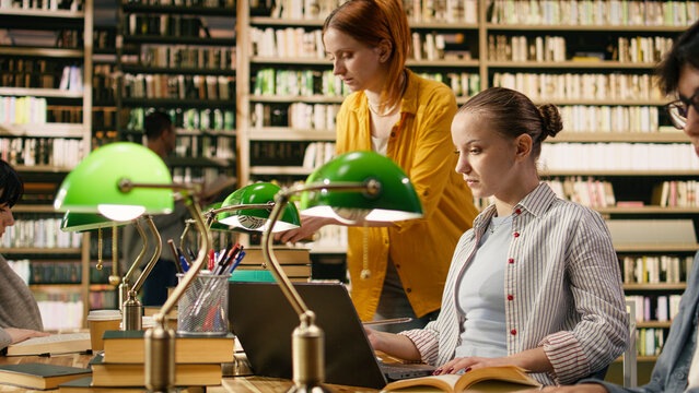 Focused student listening to classmate during library study session, surrounded by open books at wooden table together under green lamps.