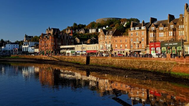 Warm light illuminates rows of Oban, Scotland seafront buildings above a stone seawall, mirrored in calm harbour water.