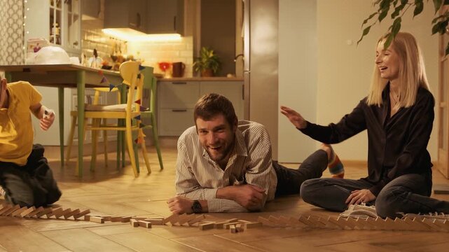 Young parents and their little boy with glasses having fun together at home, playing with a line of falling dominoes and then joyfully piling up on top of each other on the living room floor