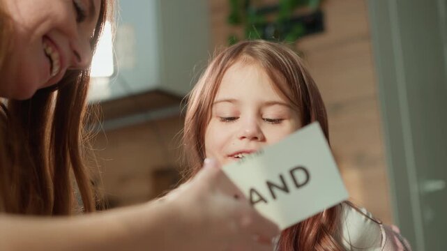 Woman interacts closely with young girl indoors helping her learn about body parts using flashcards. scene is set in home environment with soft lighting and natural elements