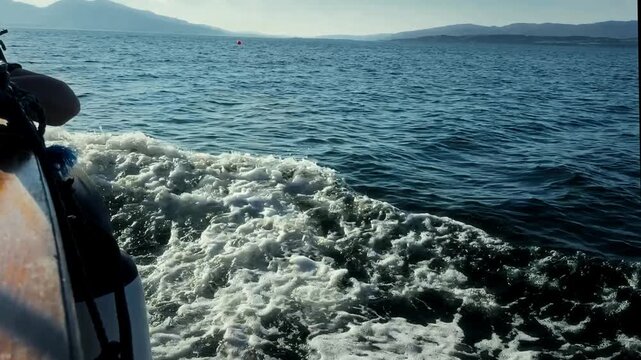 The side of a boat frames a foaming wake on blue coastal water, with distant hills near Oban, Scotland.