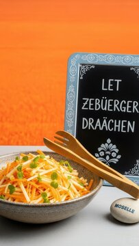 A vibrant photograph of Luxembourgish Carrot & Apple Salad with micro-mint, featuring a dramatic orange negative space backdrop and a corner detail of dried apple peel and red-white-blue linen.