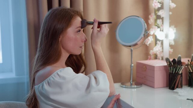 Young lady applies eyeshadow using brush in front of round mirror decorated with flowers. scene features soft lighting from nearby window and vanity lights organized makeup brushes on table