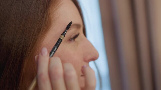 Woman applies makeup using eyebrow brush. focus remains on detailed action of her hand holding tool near her face emphasizing precision in personal care