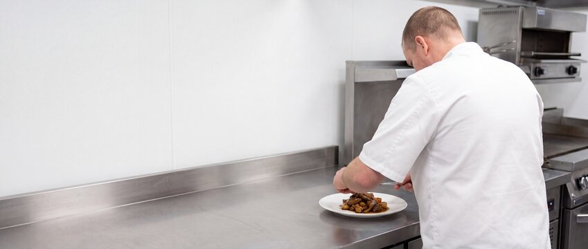 Chef plating a meat dish on a white plate in a professional stainless steel kitchen