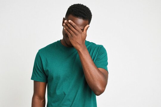 Young handsome black man wears green t-shirt on white background. Makes facepalm gesture with hand over eyes. Attractive african male shows emotion of embarrassment shame smiling. Guy feels regret