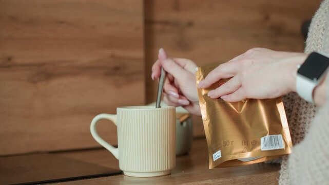 Shows two hands one holding open golden package and other using spoon to scoop granules into white ribbed mug on wooden surface. background features warm wood paneling