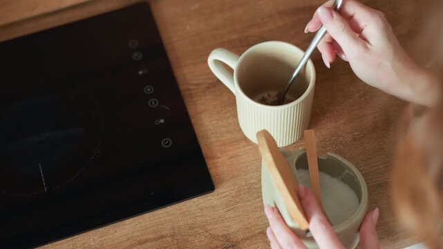 Woman hands hold and use utensils for adding sugar into cup. scene is set in domestic environment with wooden surfaces and induction cooktop visible mug